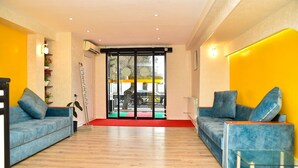 A modern hotel lobby with two blue sofas, a glass coffee table, wooden flooring, and a red carpet leading to a glass door.