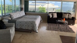 Un'ampia stanza con un letto grande, un divano scuro e un tavolino da caffè. Grandi finestre offrono vista su pannelli solari e aree verdi.