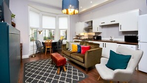 Et moderne køkken med hvide skabe, et spiseområde med bord og stole, en sofa med farverige puder og en hængende lampe.