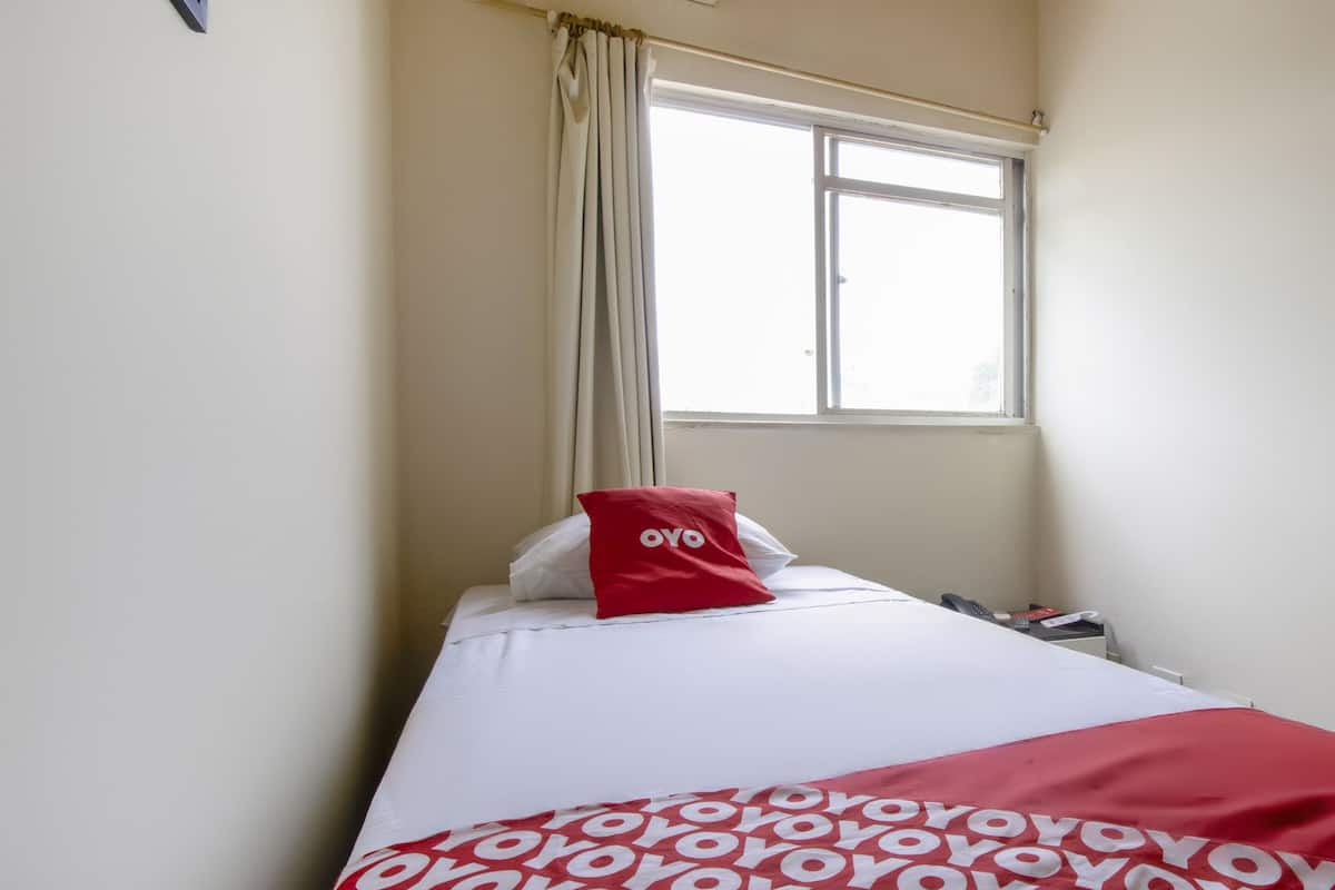 A hotel room with a bed, a window with curtains, a red pillow, and a blanket with a pattern.