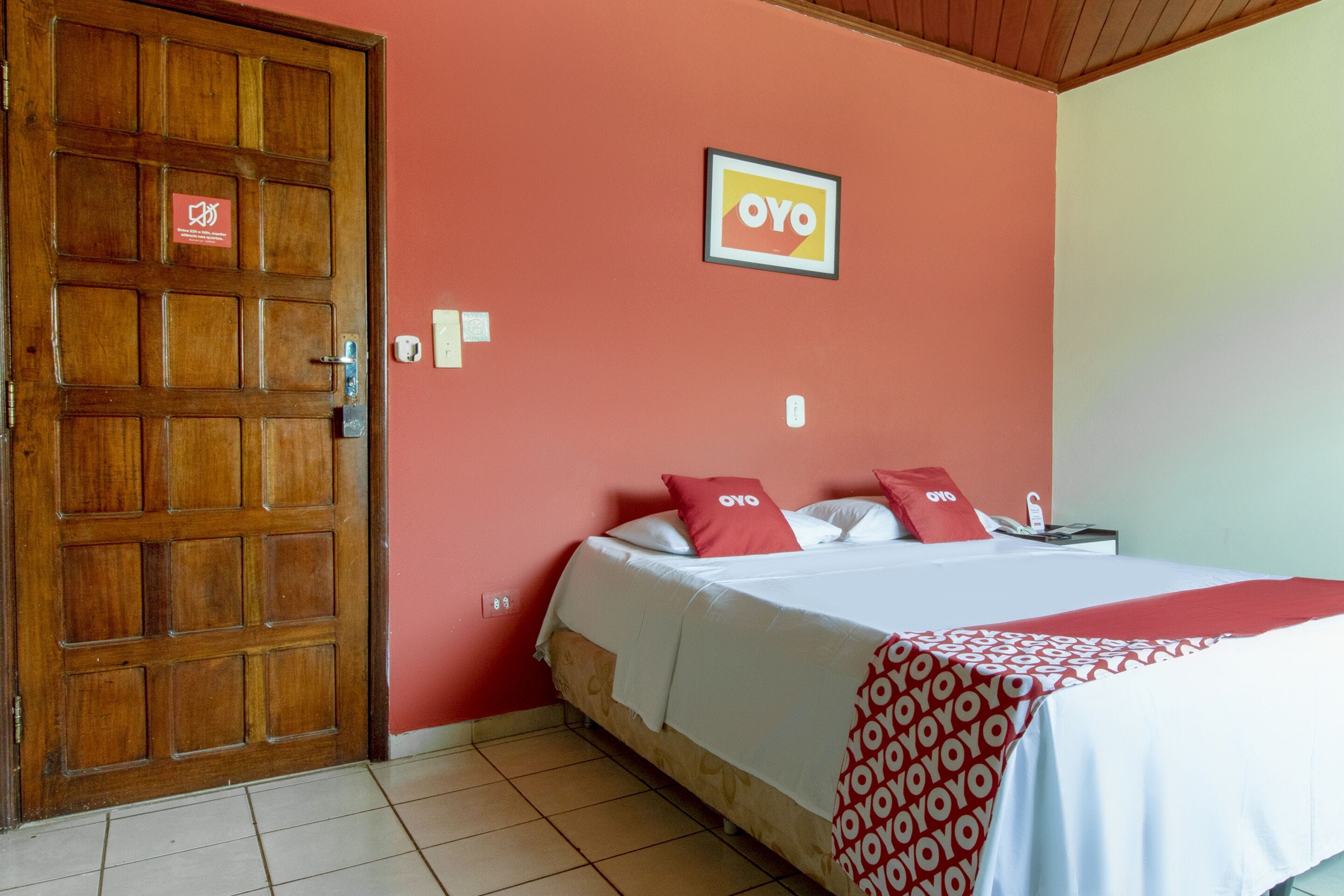 A hotel room with a wooden door, a bed with red and white bedding, and a wall-mounted sign.