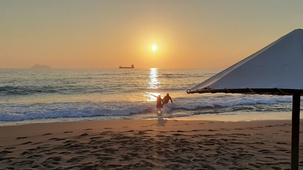 A beach scene with a boat in the water, a person standing near the water's edge, and a tent-like structure on the sand. - Kalamaki Sight Irene's (Faistos)
