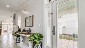 Una entrada limpia y moderna con un banco de madera, dos plantas en macetas, un espejo decorativo y una puerta de cristal que da a un patio exterior.