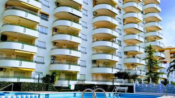 Un hotel de varios pisos con balcones, piscina y una zona ajardinada con una palmera.