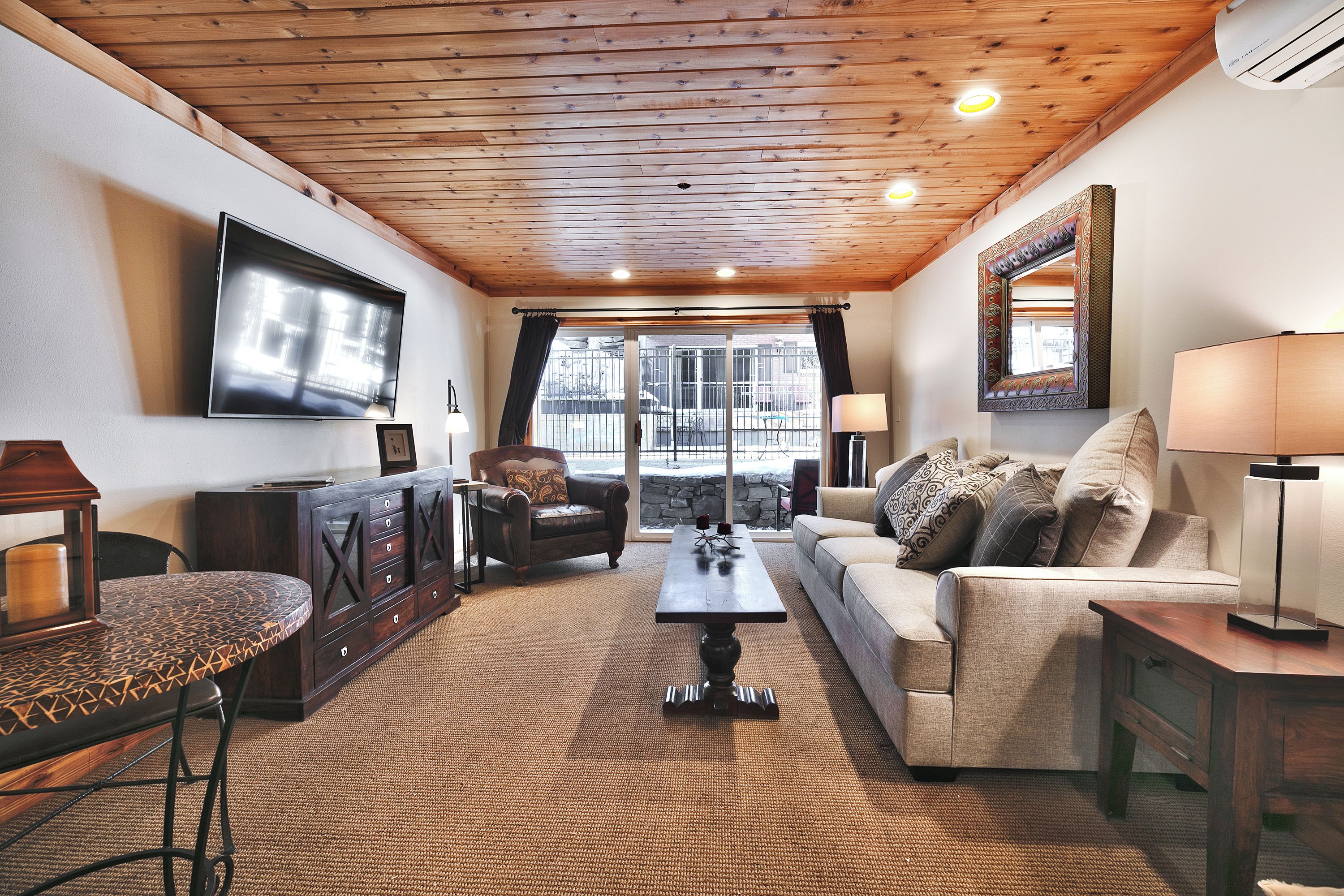 A cozy living room with a wooden ceiling, a flat-screen TV mounted on the wall, a sofa with patterned cushions, a coffee table, and a side table with a lamp.
