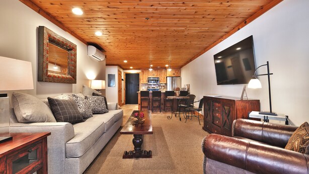A spacious living room with a wooden ceiling, a large flat-screen TV mounted on the wall, a sofa with patterned cushions, a leather armchair, and a coffee table with decorative items. - Top of Main Street - TMI with Parking (Park City)