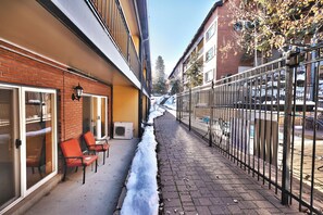 Una calle estrecha con acera nevada, un edificio de ladrillo con puerta de cristal y una valla metálica a la derecha.