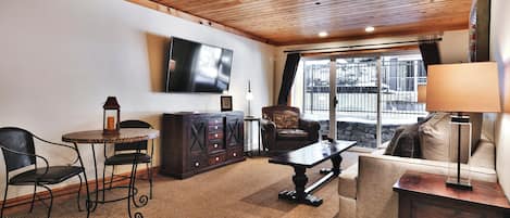 A cozy living room with a wooden ceiling, a flat-screen TV mounted on the wall, a leather armchair, a sofa, a coffee table, and a cabinet.