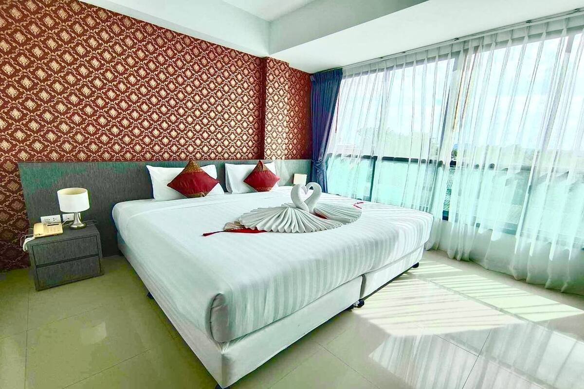 Habitación de hotel con una cama grande, una mesita de noche con lámpara, un papel tapiz estampado y vistas a través de cortinas translúcidas.