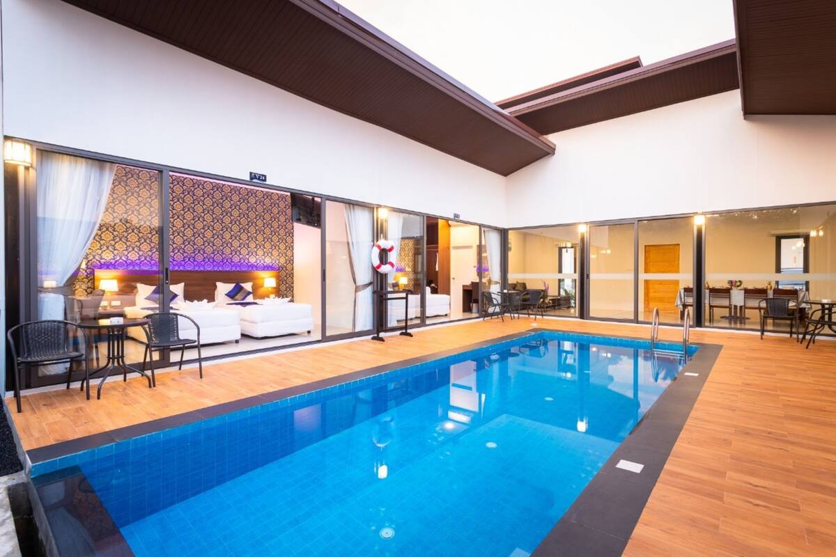 Una piscina interior con cubierta de madera, un área de estar con sofá y sillas, y vistas a una habitación con pared estampada y cama.
