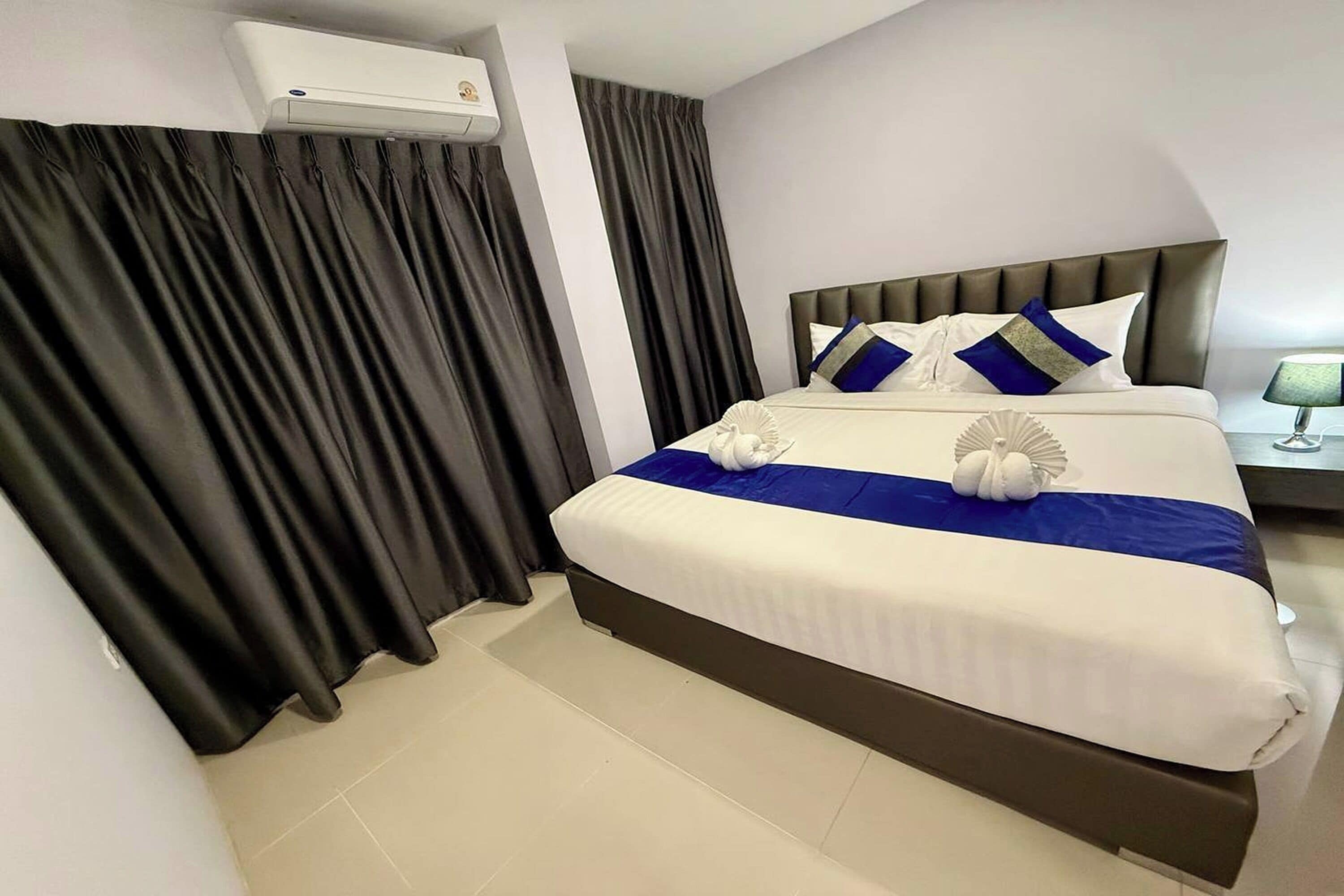 A hotel room with a large bed, dark curtains, a white air conditioning unit, and a bedside table with a lamp.