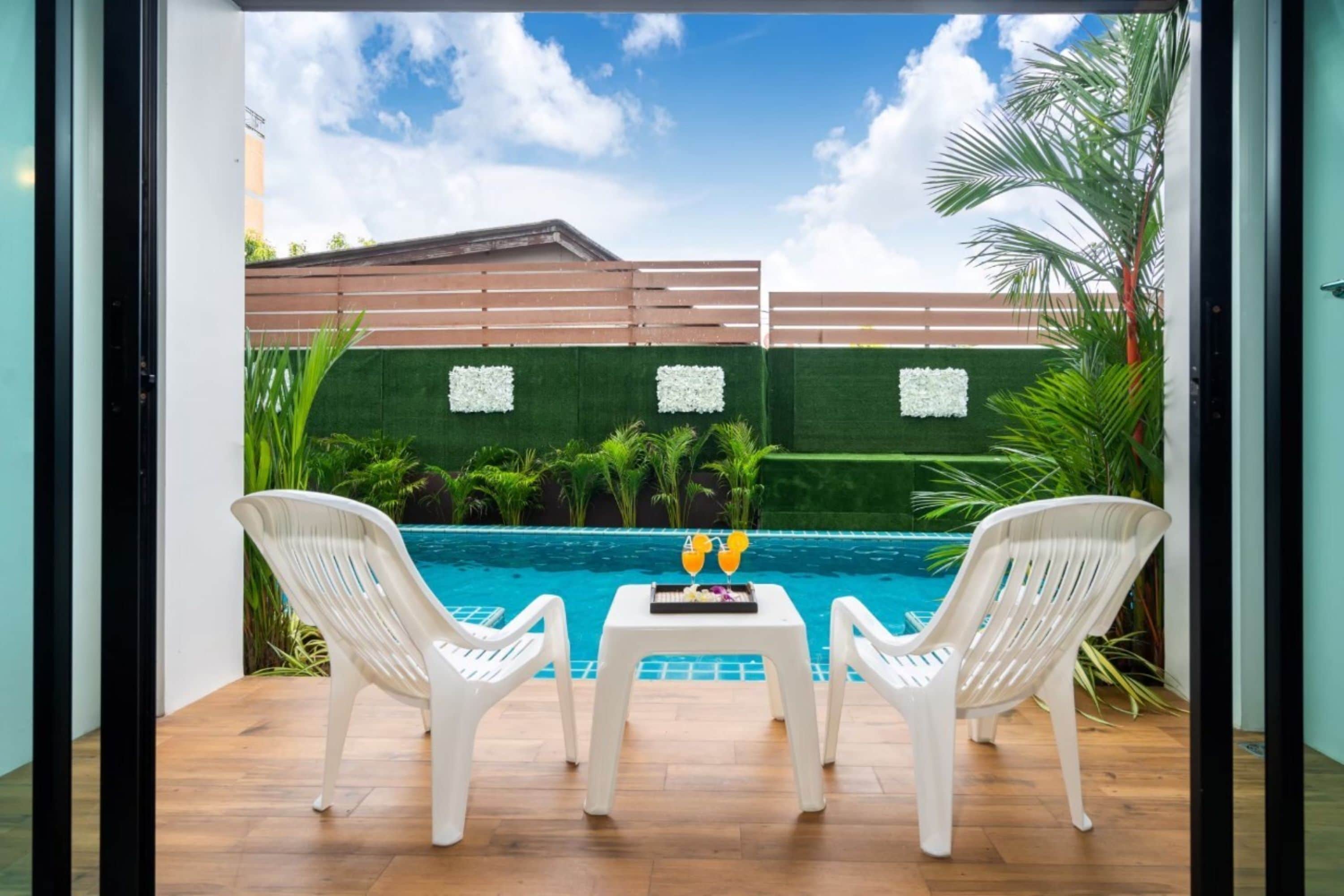 Blick auf einen Swimmingpool mit zwei weißen Plastikstühlen und einem Tisch mit Getränken, umgeben von Grünpflanzen und einem Holenzaun.