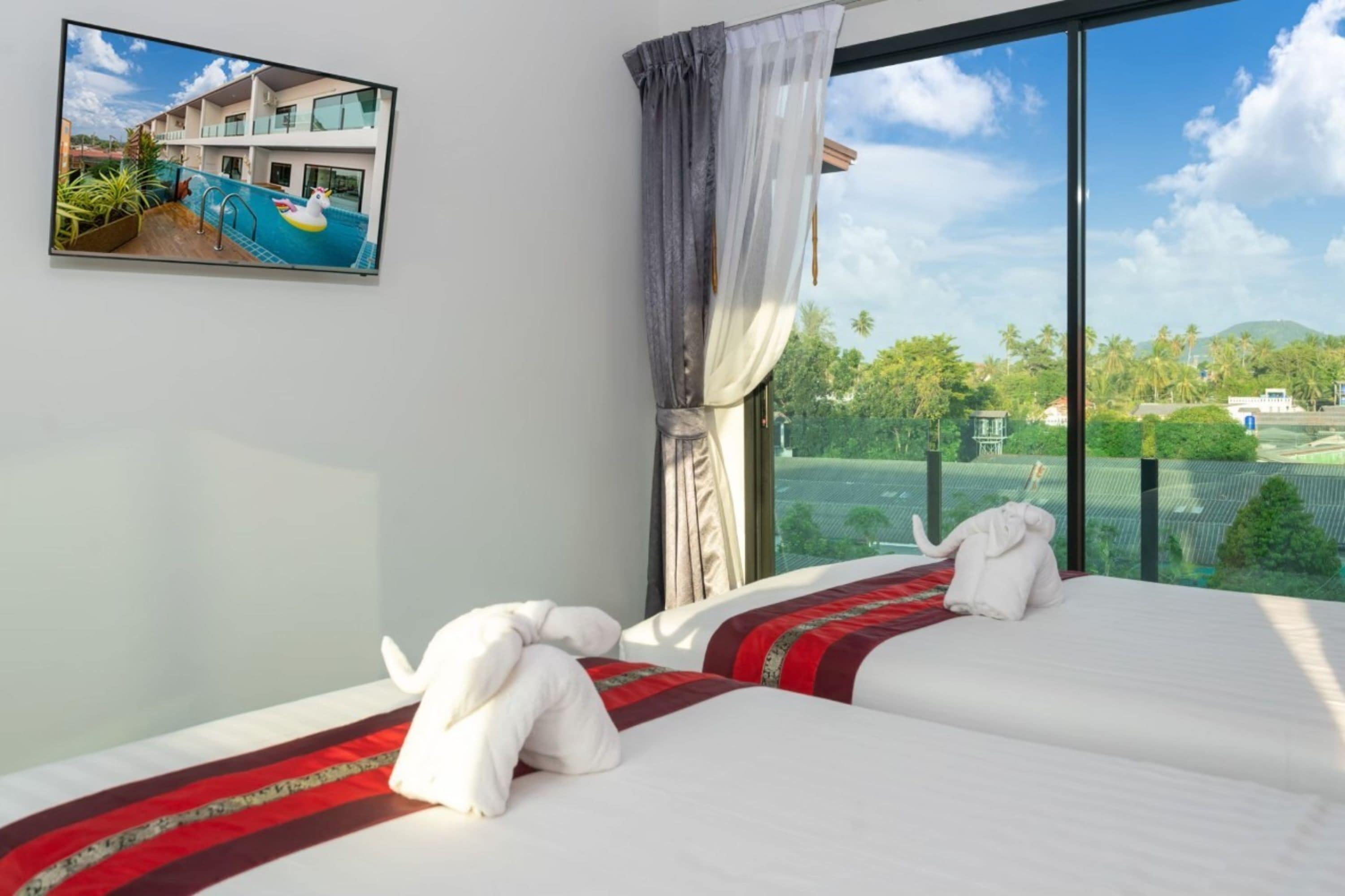 A hotel room with a large window, a bed with white linens and a red and black striped blanket, and a view of a pool and greenery outside.