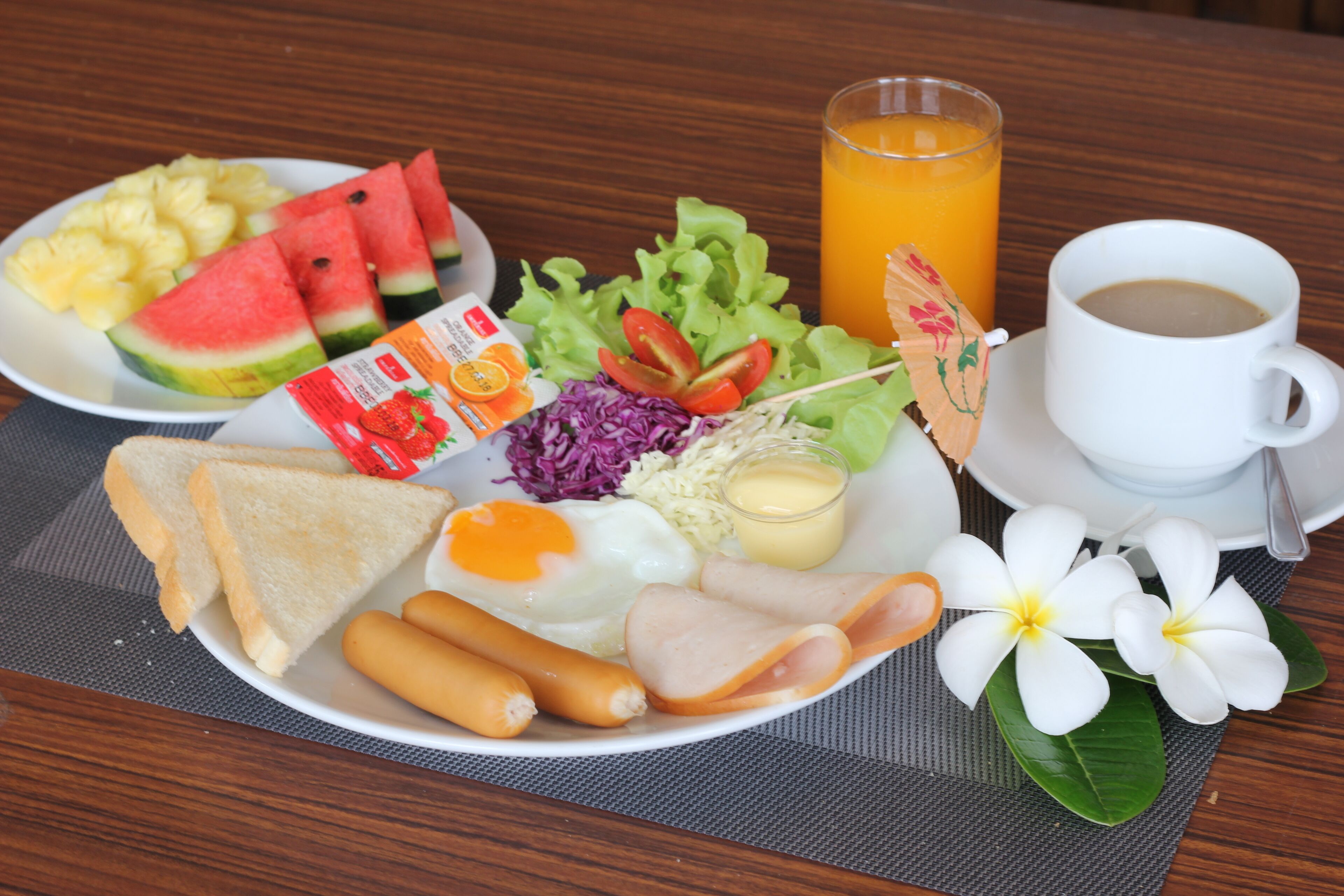 A breakfast plate with toast, eggs, ham, sausages, and vegetables, a glass of orange juice, and a cup of coffee.