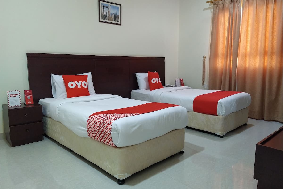 Une chambre d’hôtel avec deux lits, chacun recouvert d’une courtepointe rouge et blanche et agrémenté d’oreillers portant le logo « OYO ». On y trouve une table de chevet avec un panneau rouge et, au-dessus des lits, un tableau encadré accroché au mur.