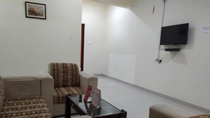 A living room with a sofa, armchair, and a glass coffee table. A flat-screen TV is mounted on the wall.