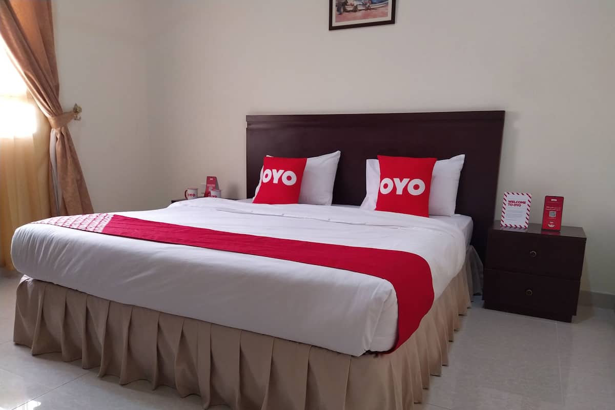 Un lit bien fait, recouvert d’une couvre-lit rouge et blanche assortie aux coussins, une table de chevet en bois avec un panneau rouge, et une base de lit beige avec une jupe.