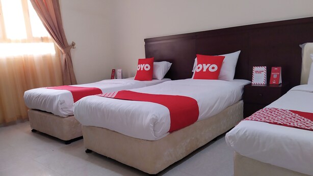 A hotel room with three beds, each with white bedding and red accents. There is a wooden headboard, a nightstand with a red sign, and a window with curtains.