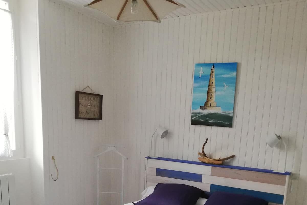 Een slaapkamer met een bed, een schilderij van een vuurtoren, een plafondventilator en een raam met gordijnen.