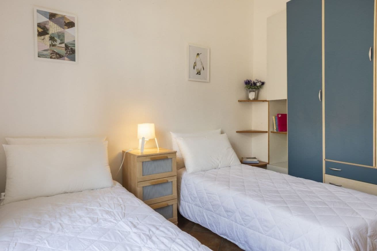 Ein Schlafzimmer mit zwei Betten, einem Nachttisch mit Lampe, einem Regal mit Büchern und einer Pflanze sowie einem Kleiderschrank.