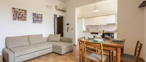 A living room with a sofa, dining table, and chairs. There is a kitchen in the background with cabinets and appliances.