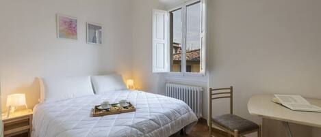 Un dormitorio con cama, mesita de noche, silla y ventana con vistas a edificios.