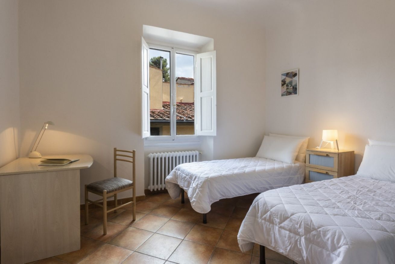 Een kleine, goed verlichte slaapkamer met twee bedden, een bureau en een stoel.