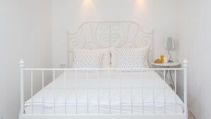 Una cama de metal blanco con un cabecero decorativo, dos almohadas, una mesita de noche con una lámpara y una pequeña mesa con un croissant y un vaso de zumo de naranja.