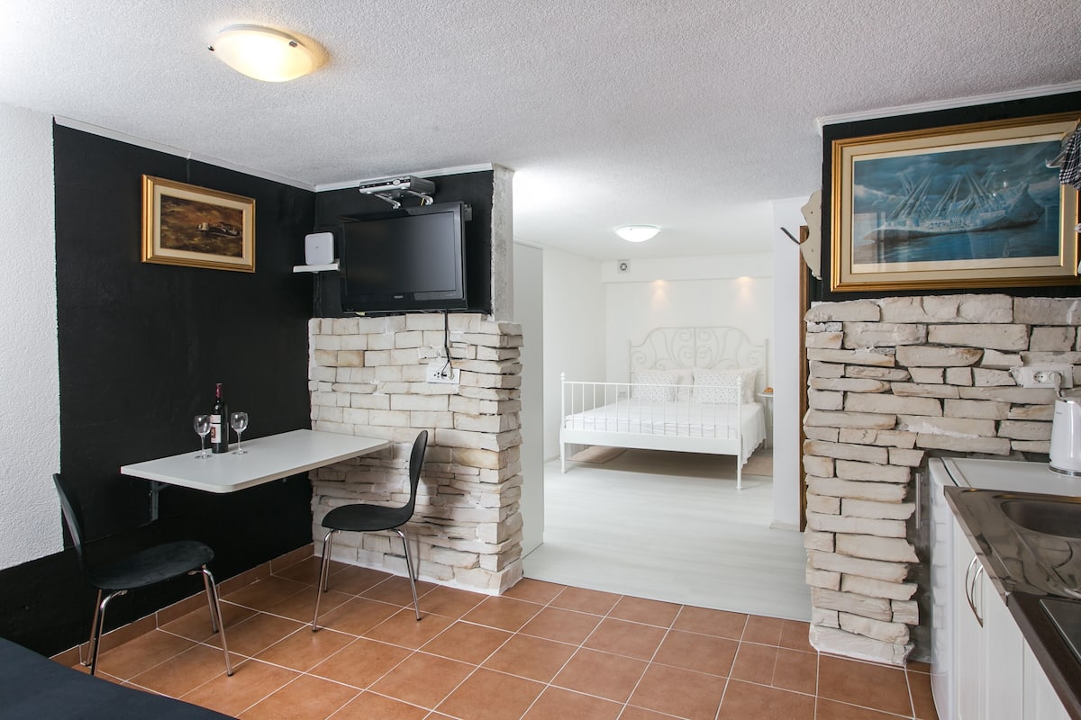 Een kleine kamer met kitchenette, een bed, een televisie bevestigd aan een stenen muur, en een eethoek met stoelen.