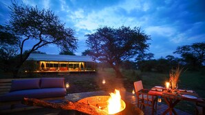 An outdoor dining area with a fire pit, wooden deck, and a view of a building in the background. - Asanja Grumeti (Serengeti)