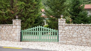 Un mur de pierre avec un portail en métal vert, entouré d’arbres et, au fond, une maison avec un toit recouvert de tuiles rouges.