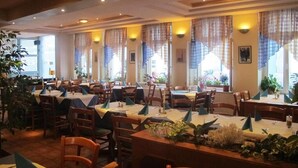A spacious dining area with multiple tables and chairs, large windows with curtains, and indoor plants. - Hotel Amadeus (Speyer)