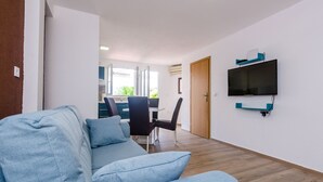 Een moderne woonkamer met een blauwe bank, een eethoek met een tafel en stoelen, en een aan de muur bevestigde televisie.