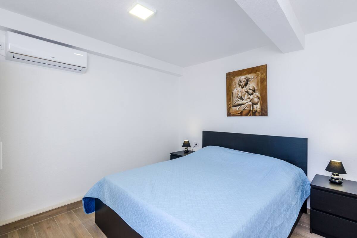 A bedroom with a bed, a nightstand, a wall-mounted air conditioner, and a painting.