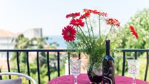 Een tafel met een fles wijn en twee glazen, een potplant met rode bloemen, en een uitzicht op groen en gebouwen op de achtergrond.