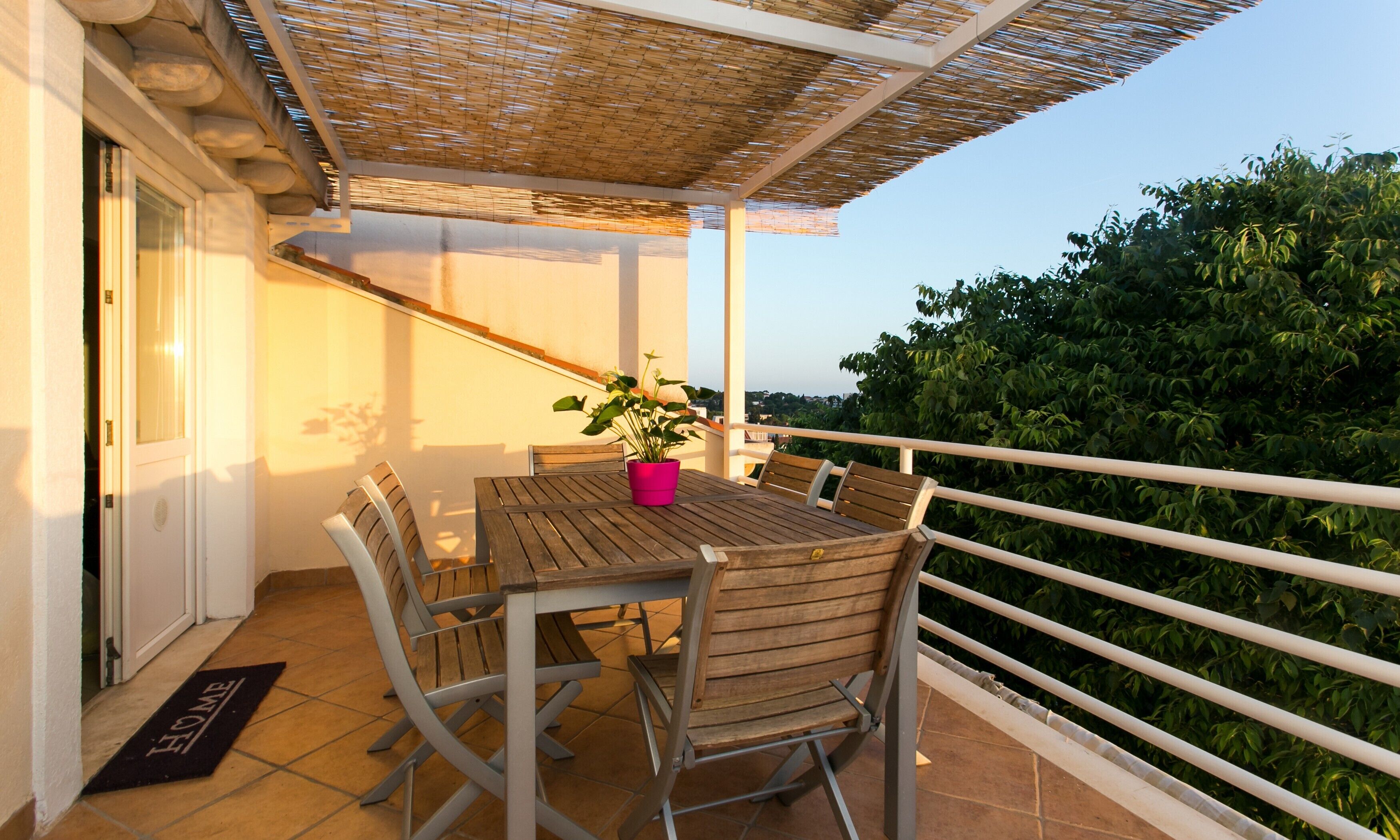 A balcony with a wooden table and chairs, a potted plant, and a view of greenery.