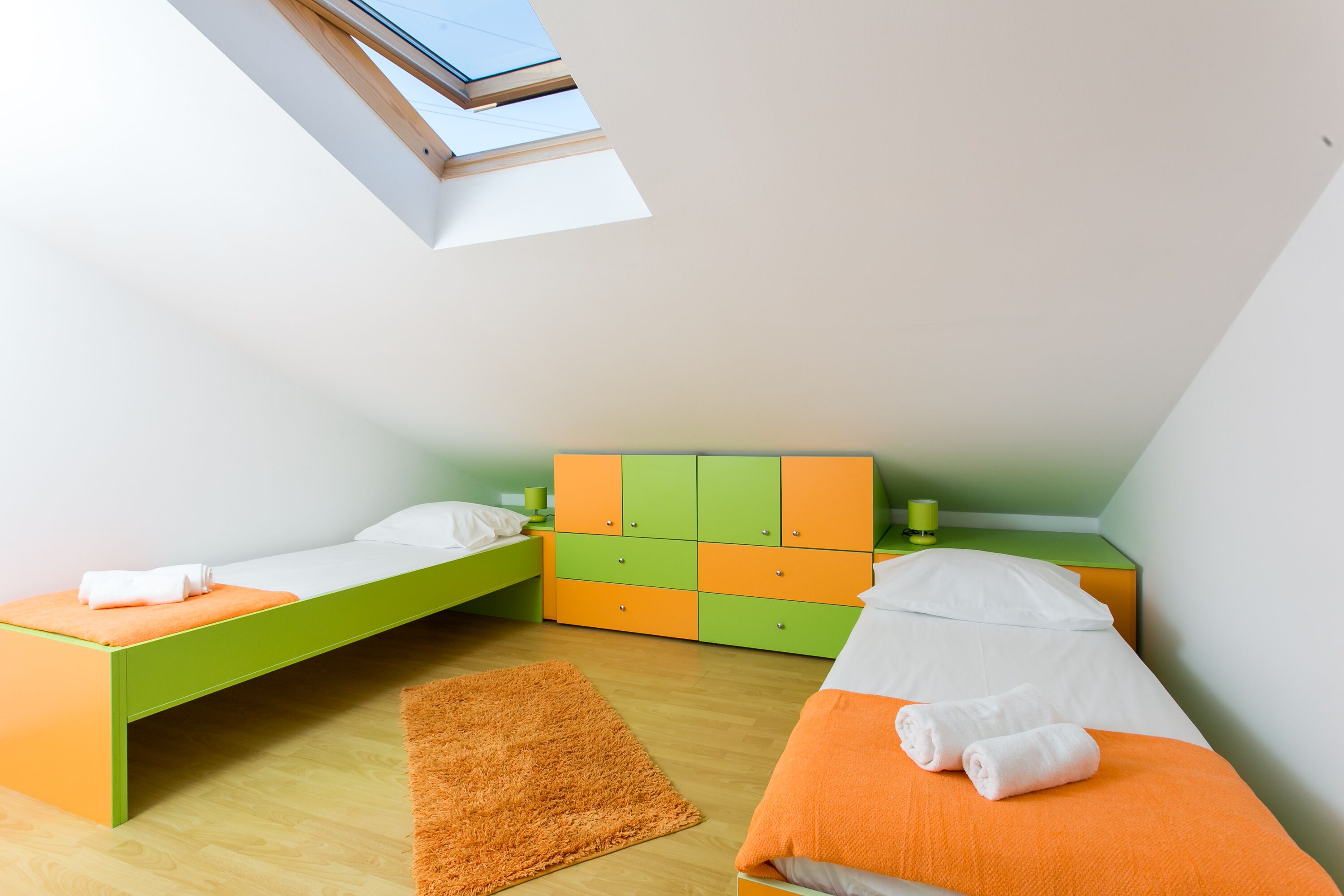 A small, brightly colored room with two beds, a green and orange dresser, and a skylight.