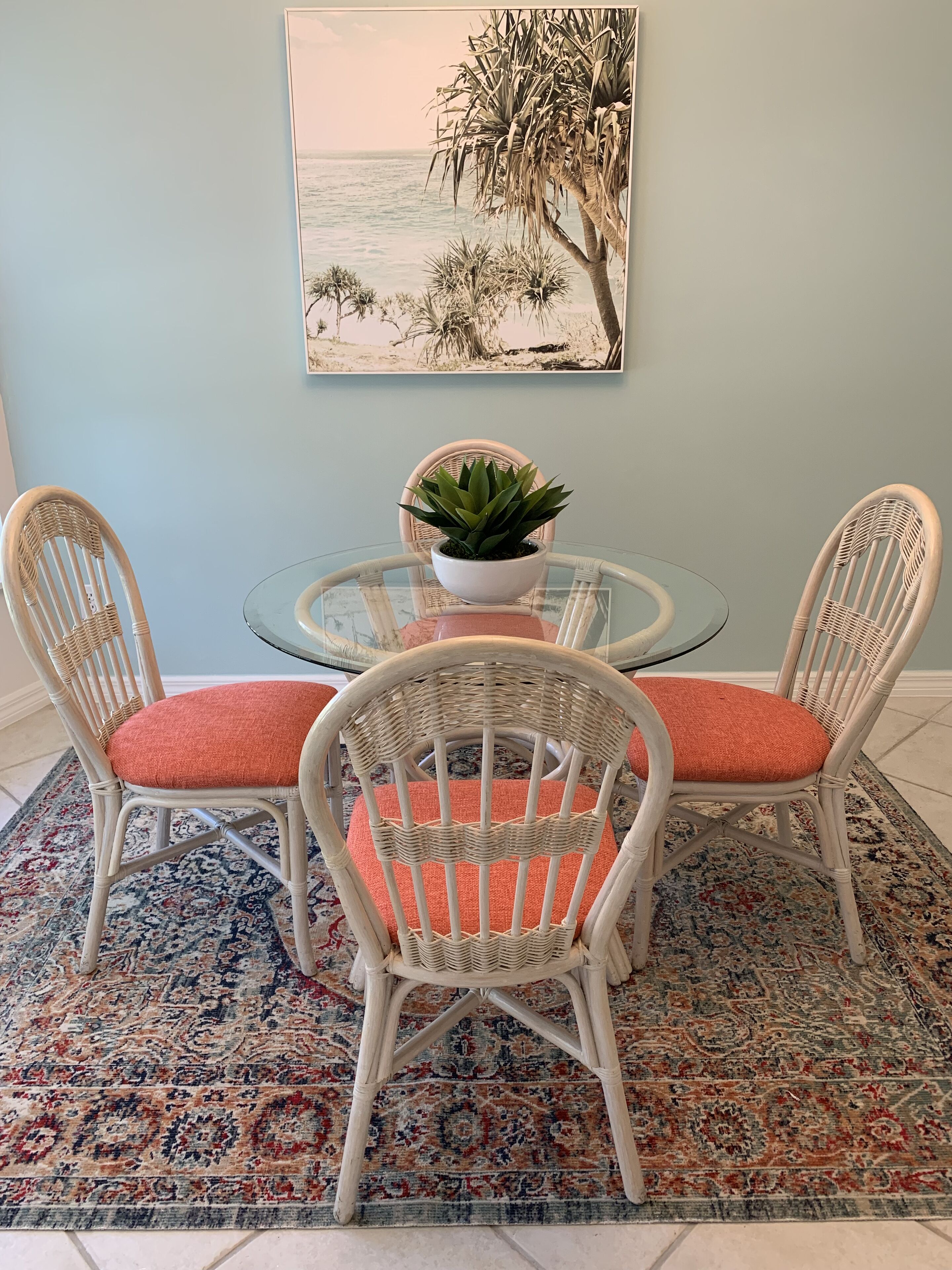 A dining area with wicker chairs and a glass table, a potted plant, and a painting of a beach scene on the wall.