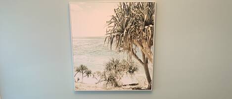 A dining area with wicker chairs and a glass table, a potted plant, and a painting of a beach scene on the wall.