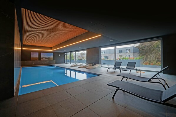 A modern indoor swimming pool with lounge chairs and a wooden ceiling design. - MY Hotel Malmedy (Malmedy)