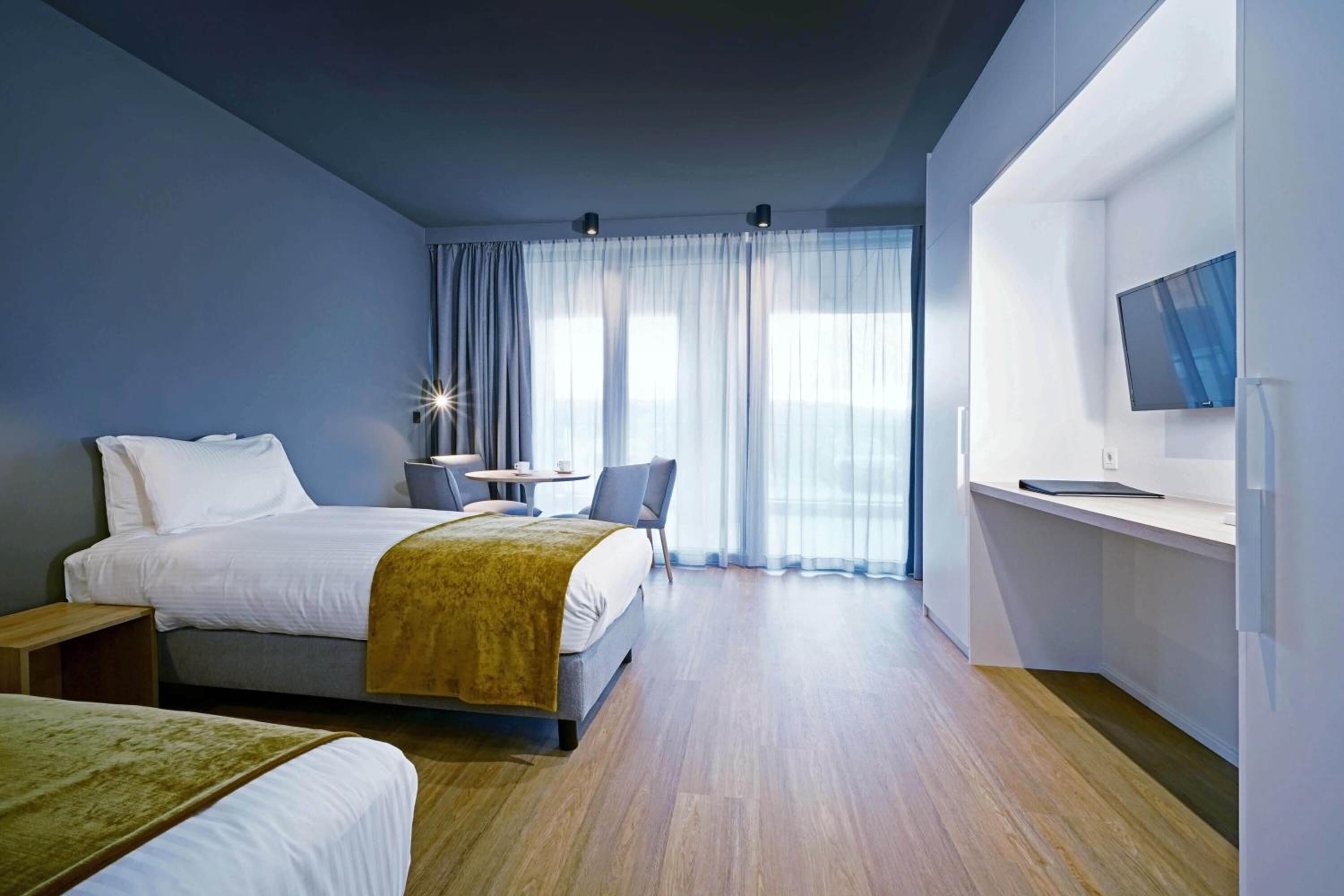 Une chambre d’hôtel moderne dotée d’un grand lit, d’un lit plus petit, d’un bureau avec une chaise, d’un téléviseur fixé au mur et d’une grande fenêtre avec des rideaux légers.