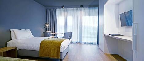 A modern hotel room with a large bed, a smaller bed, a desk with a chair, a TV mounted on the wall, and a large window with sheer curtains.