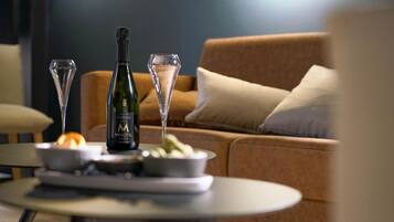 A bottle of champagne, two glasses, and a bowl of fruit on a table in a living room with a sofa and cushions.