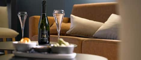 A bottle of champagne, two glasses, and a bowl of fruit on a table in a living room with a sofa and cushions.