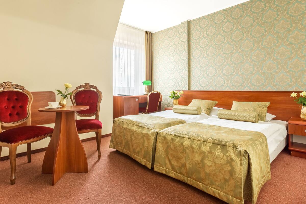 Une chambre d’hôtel avec un grand lit, deux fauteuils rouges, une table d’appoint en bois, une petite table sur laquelle est posé un vase de fleurs et une table de chevet avec une lampe.