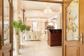 A hotel lobby with a reception desk, a large chandelier, and a dining area with tables set for a meal.