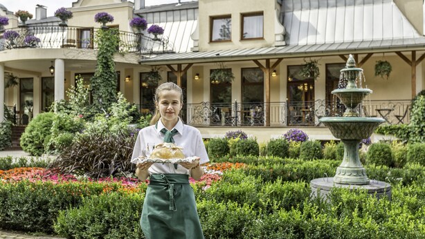 A person holding a tray with food in front of a building with a garden and fountain. - Lesna Perla - Restaurant and Hotel (Rudziniec)