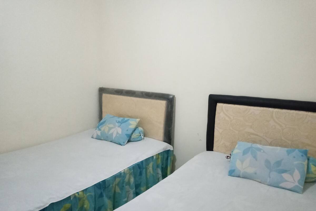 Two single beds with floral-patterned pillows and headboards, a plain wall, and a window with a curtain.