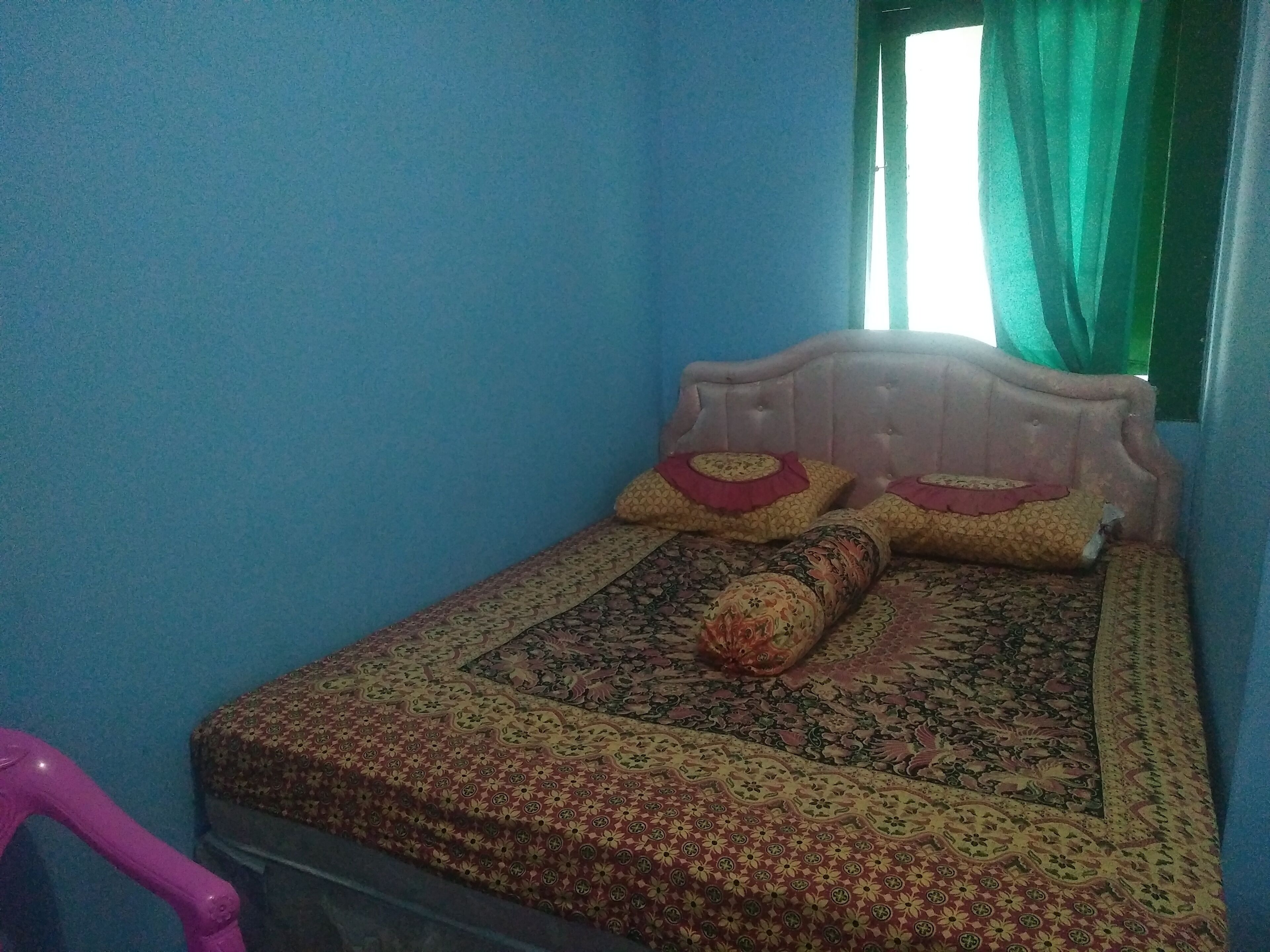 Una habitación pequeña con una cama, un ventanal con cortinas verdes y una silla rosa.
