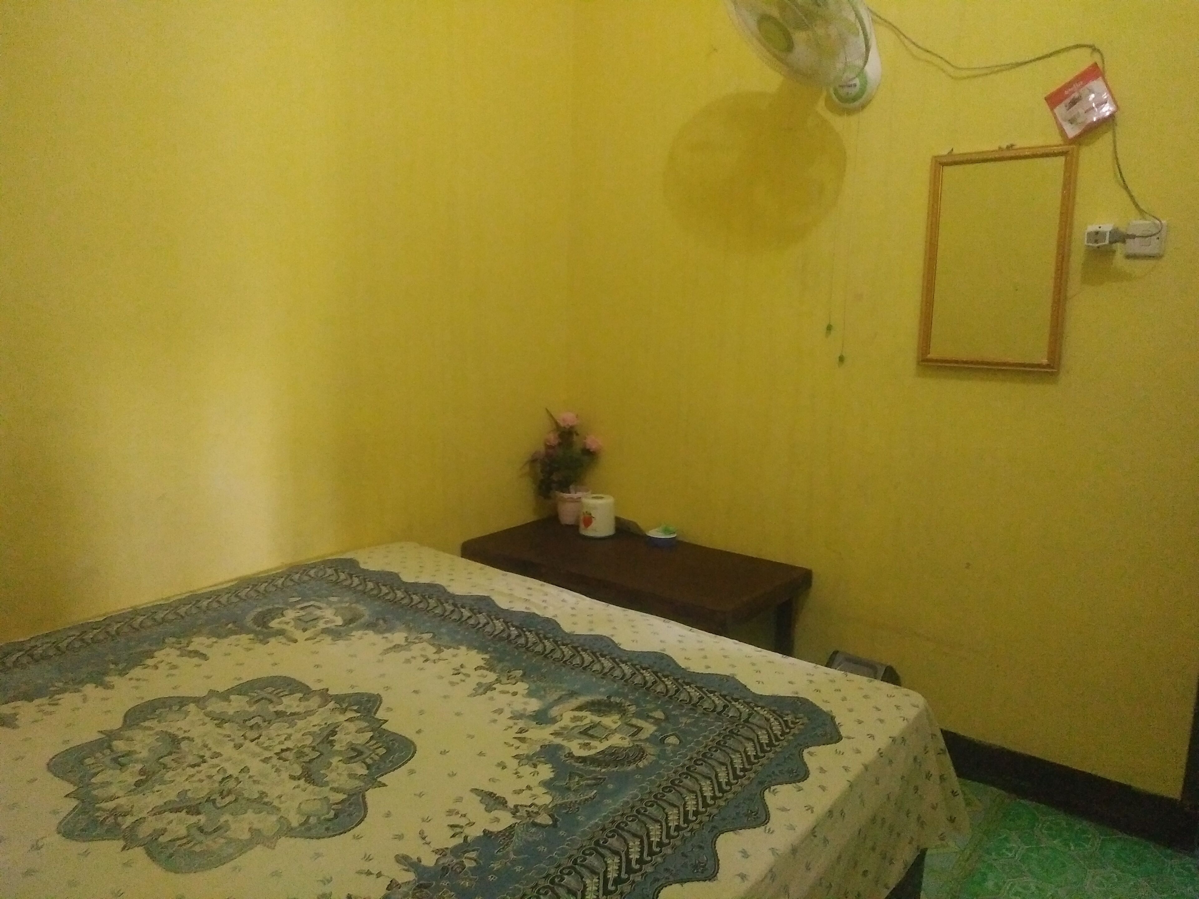 Una stanza piccola con un letto, un comodino, una ventola a muro e uno specchio.